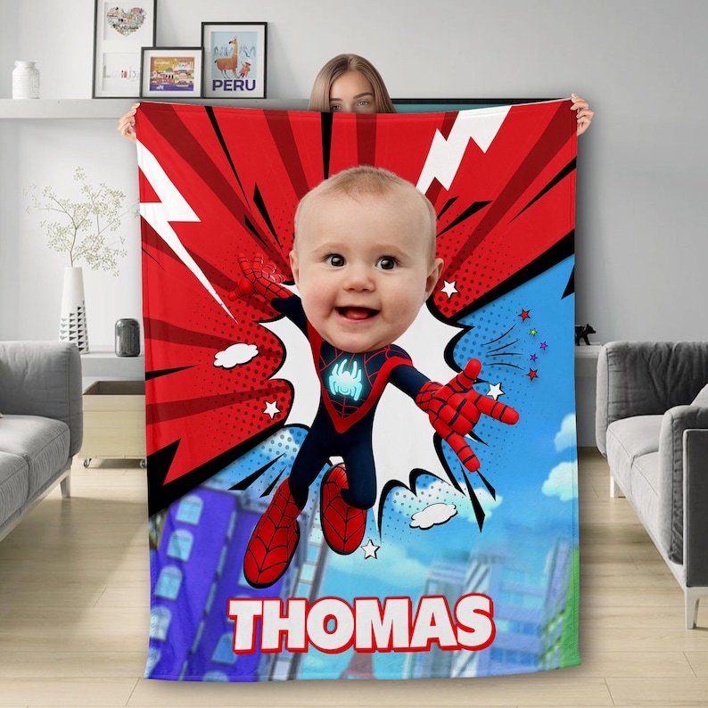 May include: A red, blue, and white fleece blanket with a cartoon superhero design. The superhero has a child's face and is wearing a black and red suit with a spider logo. The blanket has the name "THOMAS" printed on it.