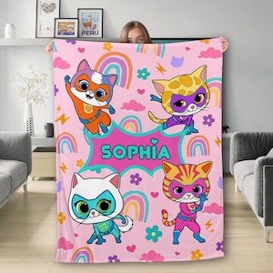 May include: Pink fleece blanket with a colorful rainbow design and four cartoon cats wearing superhero costumes. The blanket has the name "Sophia" written on it.