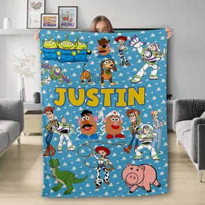 May include: A blue fleece blanket featuring a range of Toy Story characters, such as Woody, Buzz Lightyear, and Mr. and Mrs. Potato Head. The blanket has a yellow banner with the name "JUSTIN" printed on it.