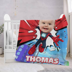 May include: A red, blue, and black personalized blanket with a comic book design featuring a baby's face in a Spiderman costume. The blanket has the name "THOMAS" printed on it.