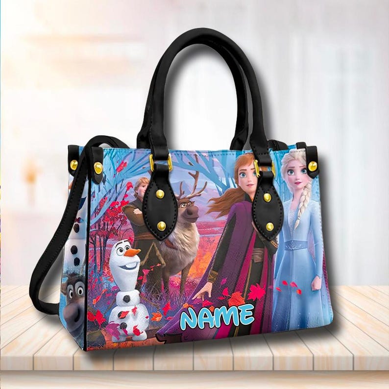 Puede incluir: Un bolso de mano azul y morado con tem&aacute;tica de Frozen. El bolso presenta im&aacute;genes de Elsa, Anna, Olaf y Sven. El bolso tiene un asa negra y una correa negra. El bolso tiene detalles dorados y la palabra "NAME" impresa en la parte delantera.