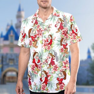 May include: A white short-sleeved button-up shirt with a tropical print featuring a cartoon character from the Disney movie Snow White and the Seven Dwarfs. The character is Grumpy, the dwarf who is always frowning. The shirt has a white background with green leaves, red flowers, and a white collar.