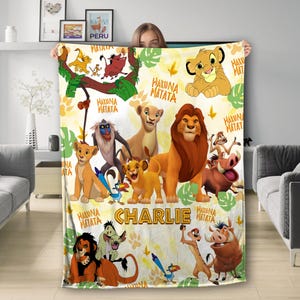 May include: A soft, yellow blanket featuring characters from The Lion King. The blanket has images of Simba, Timon, Pumbaa, and other characters, with the words "Hakuna Matata" and the name "Charlie" printed on it.