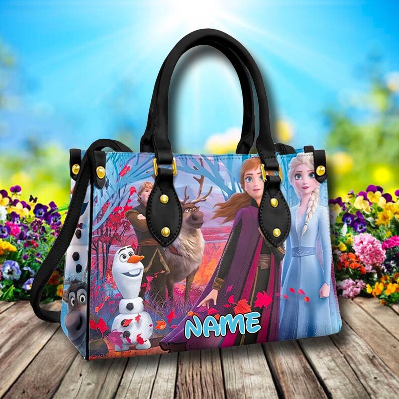 Puede incluir: Un bolso de mano negro con un estampado azul, blanco y morado con personajes de la pel&iacute;cula Frozen. El bolso tiene detalles dorados y el texto "NAME" est&aacute; impreso en la parte delantera.