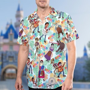 May include: A white short-sleeved button-down shirt with a colorful print of characters from the Disney Pixar movie Coco. The print features Miguel, Hector, Dante, and other characters from the film. The shirt has a tropical-inspired design with palm leaves and pineapples.