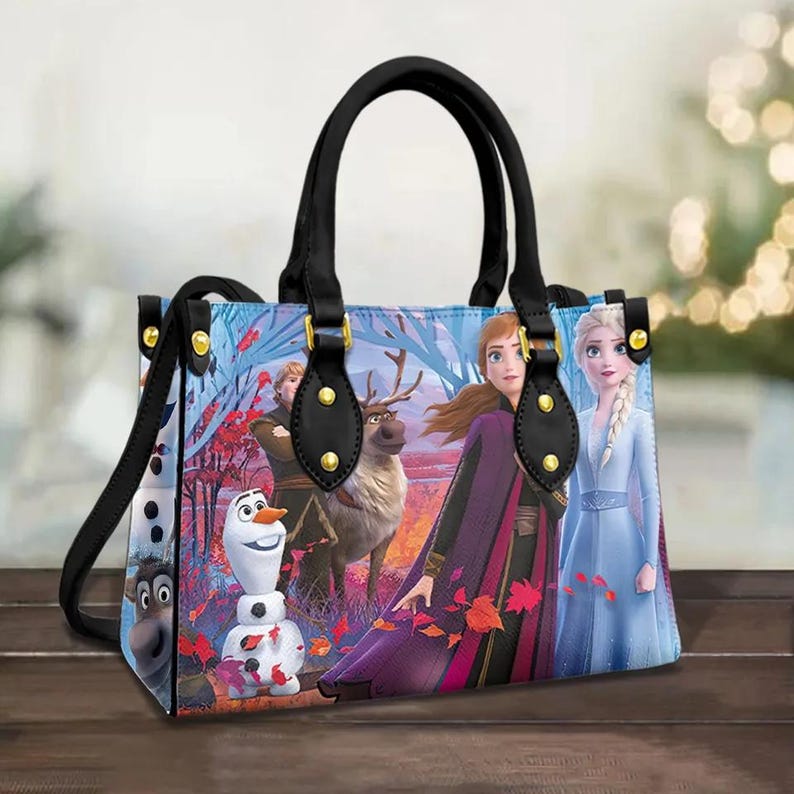 Puede incluir: Un bolso de mano de cuero negro con detalles dorados y un estampado de personajes de la pel&iacute;cula Frozen. El estampado presenta a Elsa, Anna, Olaf y Kristoff en un paisaje invernal nevado.