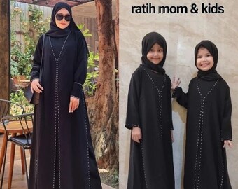 May include: Three people wearing black abayas with beaded trim. The woman is wearing a black headscarf and sunglasses. The two children are wearing black headscarves. The text "ratih mom & kids" is in the top right corner of the image.