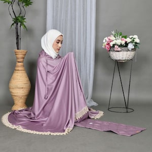 May include: A woman wearing a purple prayer shawl with a white lace trim and a white headscarf. She is kneeling on a purple prayer rug with a white lace trim.