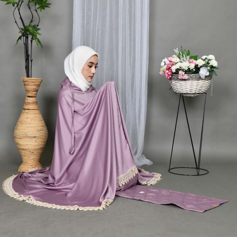 May include: A woman wearing a purple prayer shawl with a white lace trim and a white headscarf. She is kneeling on a purple prayer rug with a white lace trim.