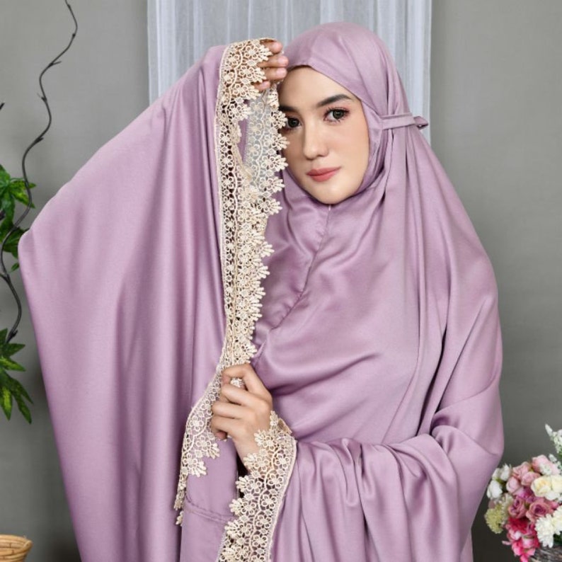May include: A lavender-colored prayer shawl with delicate white lace trim. The shawl is draped over a person's shoulders and head.