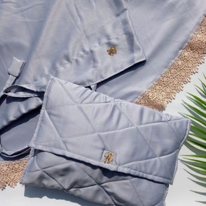 May include: A gray quilted prayer rug with a matching gray carrying case. The rug has a delicate lace trim and a gold embroidered design. The case has a gold embroidered initial.