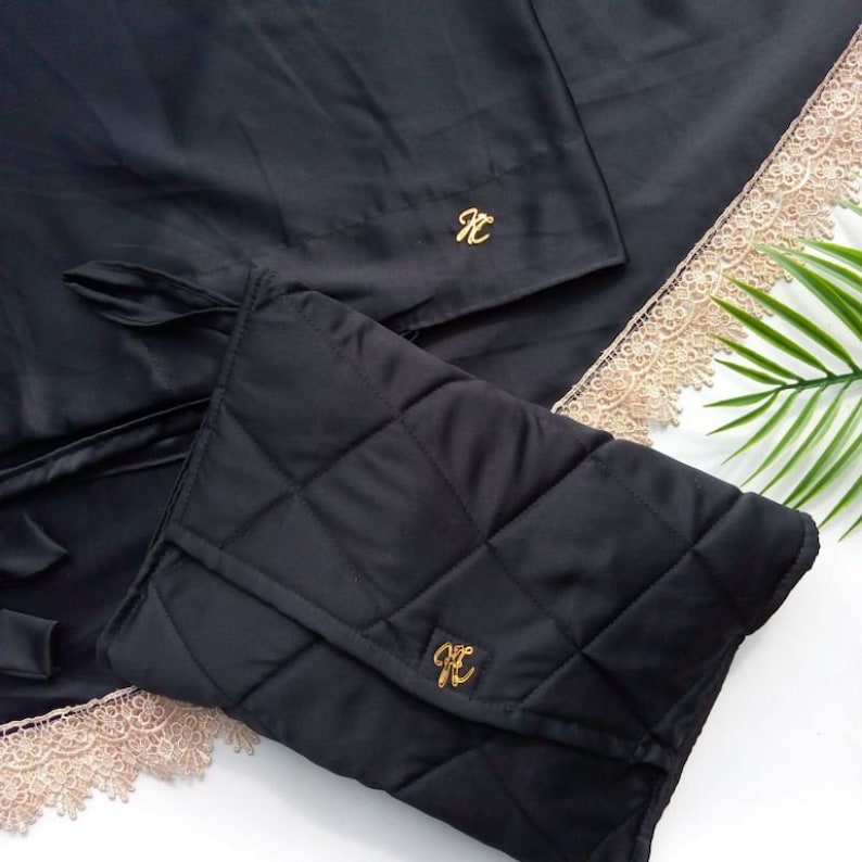 May include: Black quilted prayer rug with a gold embroidered initial "K" on the front and a matching black satin prayer rug with a gold embroidered initial "K" on the top right corner. Both rugs have a beige lace trim.