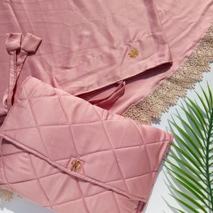 May include: A pink quilted prayer rug with a matching pink satin cover and a gold embroidered logo. The rug has a lace trim and is folded in half.