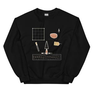 May include: A black crewneck sweatshirt featuring a white graphic print of archaeological tools including a grid, a trowel, a brush, a tag, and a cross-section of soil layers. The design is centred on the chest and uses simple line art.