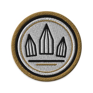 May include: A circular embroidered patch featuring a light gray center with three black-outlined pencil shapes pointing upward. The design is framed by a thick black border and an outer edge of metallic gold thread, with two horizontal lines below.