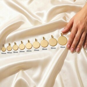 A hand is holding a row of gold-colored pendants, each with a different size, against a white fabric background.