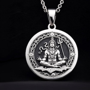May include: A silver pendant necklace featuring a detailed depiction of a seated deity in a meditative pose. The pendant has an ornate border and is suspended from a silver chain. The deity holds a trident and is surrounded by a snake.