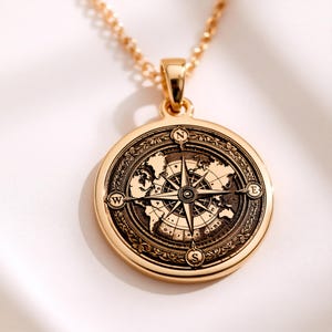 Puede incluir: Collar con colgante dorado con un diseño detallado de rosa de los vientos y un fondo de mapa del mundo. El colgante redondo cuelga de una delicada cadena. La rosa de los vientos tiene marcadores direccionales y un borde floral detallado.