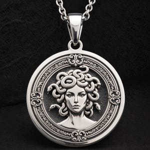 Op de afbeelding: Een zilveren hangerketting met een afbeelding van Medusa. De ronde hanger toont een gedetailleerde afbeelding van Medusa's gezicht, met slangen als haar, omgeven door een sierlijke rand. De ketting heeft een zilveren ketting.