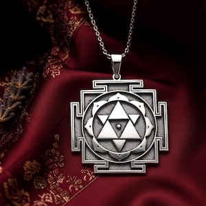 Könnte beinhalten: Eine silberne Halskette mit Anhänger, die ein geometrisches Design mit einem zentralen Dreieck und kreisförmigen Mustern aufweist. Der Anhänger hängt an einer zarten Kette vor einem tiefroten Stoffhintergrund.