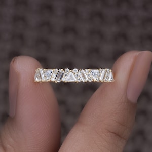 May include: A gold ring featuring a series of white, geometric-shaped stones. The ring has a mix of rectangular, triangular, and round-cut stones, creating a unique design. The ring is held between two fingers against a blurred background.