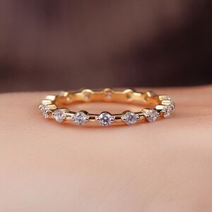 May include: A gold band ring with a continuous row of small, clear gemstones. The ring is set against a blurred background, highlighting the ring's design. The ring appears to be made of gold.