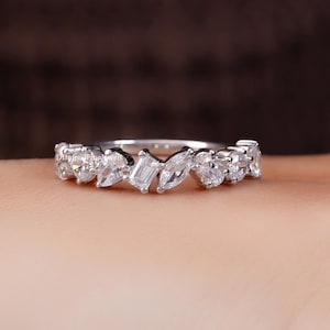 May include: A silver-toned ring featuring a row of clear, multi-shaped stones including round, marquise, and baguette cuts. The band is polished and metallic, showcasing a variety of faceted gems arranged horizontally across the top of the ring.