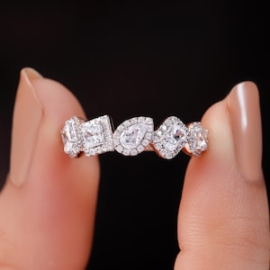 May include: A diamond ring featuring a series of differently shaped stones, including square, pear, and emerald cuts, each surrounded by a halo of smaller diamonds. The ring is held against a black background.