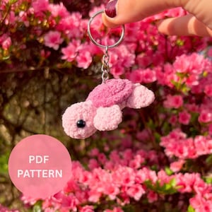 Puede incluir: Un llavero de tortuga de crochet rosa con ojos negros. El llavero está colgado de una cadena y un anillo plateados. La tortuga está enfocada y el fondo está desenfocado.