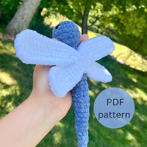 Puede incluir: Un juguete de libélula de crochet hecho con hilo azul y blanco. La libélula tiene un cuerpo largo y delgado y dos alas grandes. El juguete está siendo sostenido en una mano. El texto "PDF pattern" es visible en la imagen.
