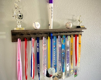 May include: A dark wooden medal display shelf with a variety of sports medals hanging below. Above the shelf are a baseball, a soccer trophy, and a white tube with "MVP" printed on it.