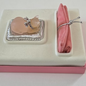 Peut inclure: Une boîte à bijoux décorative avec une figurine de chat rose sur un coussin argenté. La boîte a un dessus blanc et une base rose. Un tissu rose est maintenu par un anneau argenté sur le côté droit.