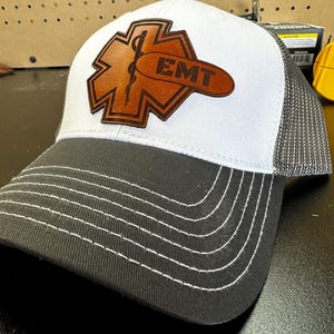May include: White and grey trucker hat with a brown leather patch featuring a medical symbol and the word "EMT".