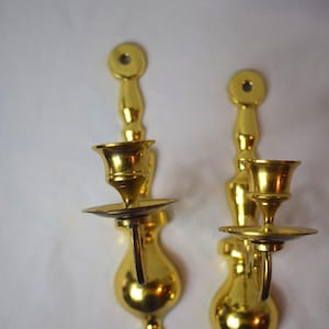 May include: Two gold-colored wall sconces. Each sconce has a round top with a hole, a long stem, a candle holder, and a small dish. The sconces are made of a shiny, reflective material.