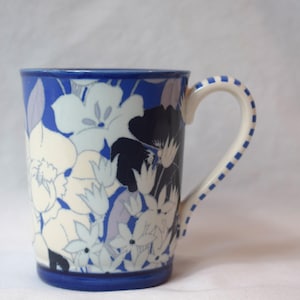 May include: A ceramic mug with a blue rim and handle, adorned with a floral pattern in white, navy, and lavender. The handle is white with blue square accents. The mug is designed for hot beverages.