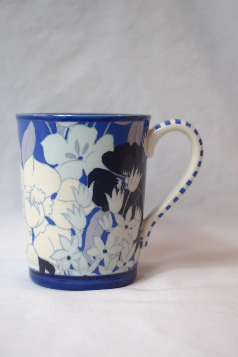 May include: A ceramic mug with a blue rim and handle, adorned with a floral pattern in white, navy, and lavender. The handle is white with blue square accents. The mug is designed for hot beverages.