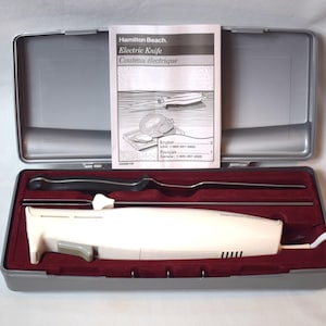 Puede incluir: Un cuchillo eléctrico blanco con una funda de transporte roja y blanca. El cuchillo está en la funda con el cable enchufado. La funda tiene una etiqueta que dice "Hamilton Beach Electric Knife Couteau electrique".