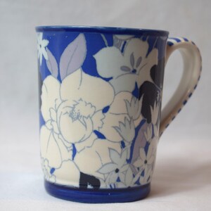 May include: A ceramic mug with a blue rim and handle, adorned with a floral pattern in white, light blue, and lavender hues. The mug's body features large, stylized flowers against a blue background, creating a decorative design.