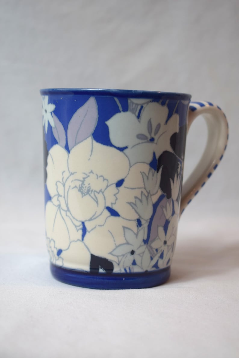 May include: A ceramic mug with a blue rim and handle, adorned with a floral pattern in white, light blue, and lavender hues. The mug's body features large, stylized flowers against a blue background, creating a decorative design.