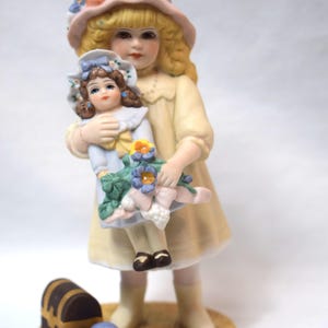 Könnte beinhalten: Eine Porzellanfigur eines jungen Mädchens in einem rosa Hut und einem gelben Kleid, das eine kleinere Puppe mit einem blauen Kleid und Blumen hält. Die Figur steht auf einem Holzsockel mit einer kleinen Truhe und einem blauen Ball.