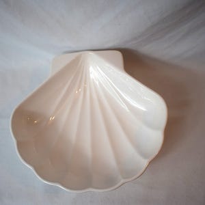 Könnte beinhalten: Eine glänzende, cremeweiße Keramikschale in Form einer großen Jakobsmuschel. Die Schale weist eine Reihe erhabener, strahlenförmiger Rippen auf, die vom flachen Scharnierbereich zum gewellten, geschwungenen Rand verlaufen und ein Becken bilden.