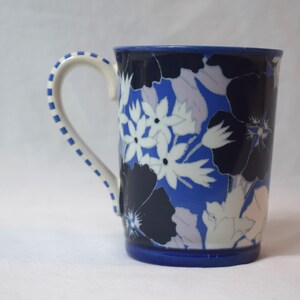 May include: A ceramic mug with a blue base and rim, featuring a floral pattern in white, navy, and light blue. The handle is white with blue dotted accents. The mug is designed for hot beverages.