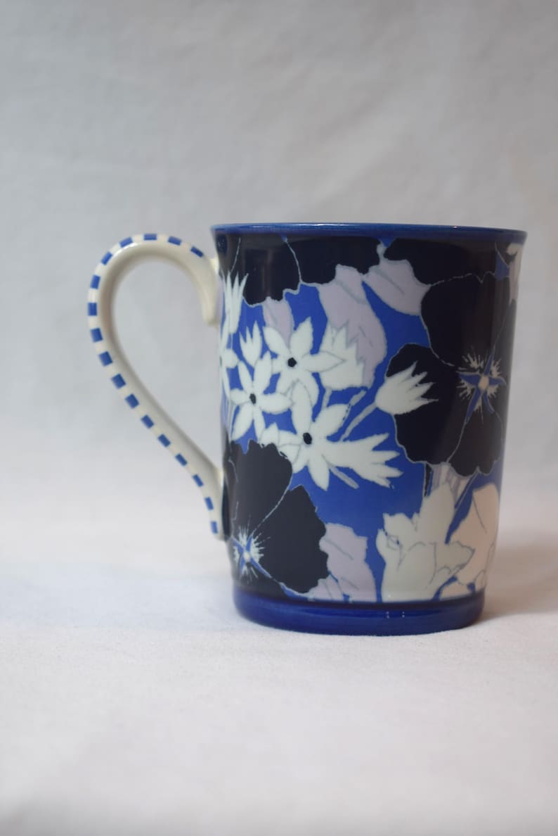 May include: A ceramic mug with a blue base and rim, featuring a floral pattern in white, navy, and light blue. The handle is white with blue dotted accents. The mug is designed for hot beverages.