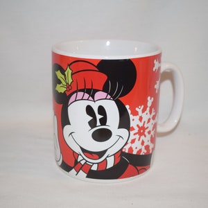 May include: A red ceramic mug with a white handle features a cartoon image of Minnie Mouse wearing a red hat with a green holly sprig and a black and red scarf. The background is red with white snowflakes.