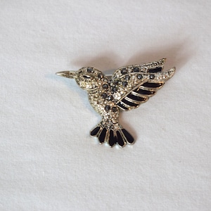 May include: A silver-toned hummingbird brooch with a detailed design. The bird's body is encrusted with small, clear crystals, and its wings and tail feature black enamel accents. The brooch is set against a white background.