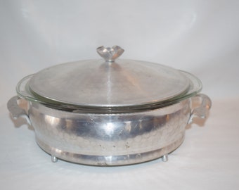 May include: A silver aluminum serving dish with a glass lid and two handles. The dish has a hammered texture and sits on three small feet.