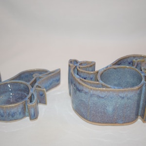 Puede incluir: Dos portavelas de cerámica con forma de pájaro con un esmalte azul y marrón. Los portavelas tienen una pequeña hendidura redonda en el centro para una vela.