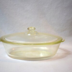 May include: A clear, oval-shaped glass casserole dish with a matching lid. The dish has a slightly yellow tint and small handles on either side. The lid has a small knob for easy handling. This vintage cookware is perfect for baking and serving.