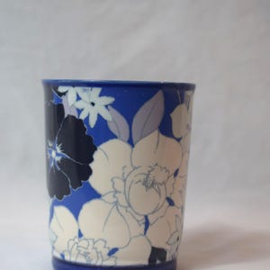 May include: A cylindrical ceramic cup with a blue base and rim, adorned with a floral pattern in white, navy, and light blue. The flowers are stylized and cover the surface of the cup, creating a decorative design.