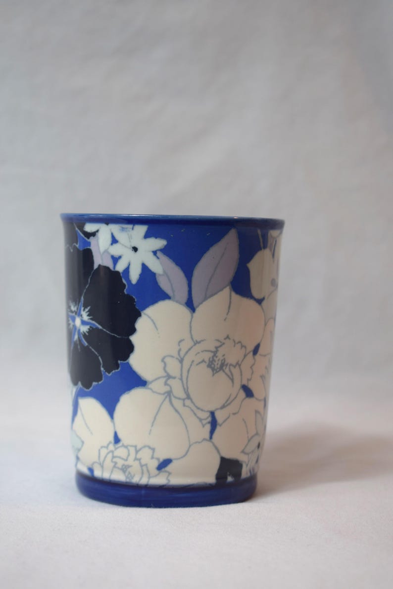 May include: A cylindrical ceramic cup with a blue base and rim, adorned with a floral pattern in white, navy, and light blue. The flowers are stylized and cover the surface of the cup, creating a decorative design.